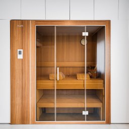 DREAMS DESIGN STUDIO - Nowoczesna sauna z drewnianym wykończeniem i szklanymi drzwiami, wkomponowana w minimalistyczne wnętrze z białymi szafkami i zasłonami.
