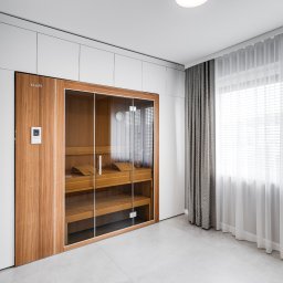 DREAMS DESIGN STUDIO - Nowoczesna sauna z przeszklonymi drzwiami w jasnym wnętrzu. Drewniane ławy, minimalistyczny design, subtelne oświetlenie i białe zasłony w oknie.