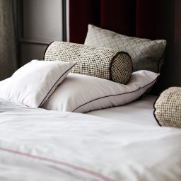 DREAMS DESIGN STUDIO - Elegancka aranżacja łóżka z białymi poduszkami z bordową lamówką, dekoracyjnym wałkiem i poduszką w kratę na tle bordowego zagłówka i zasłony.