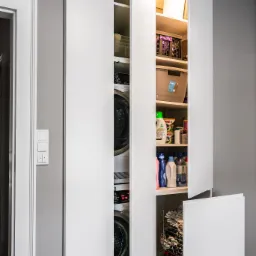 Nowoczesna szafa wnękowa z ukrytą pralką, suszarką i półkami na środki czystości. Minimalistyczny design, białe fronty, szare ściany, oświetlenie LED wewnątrz.