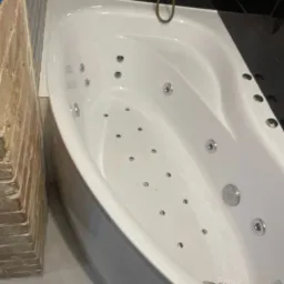 Biała wanna z hydromasażem w narożniku łazienki wykończonej czarną glazurą, widoczny fragment ceglanej ściany obok wanny.