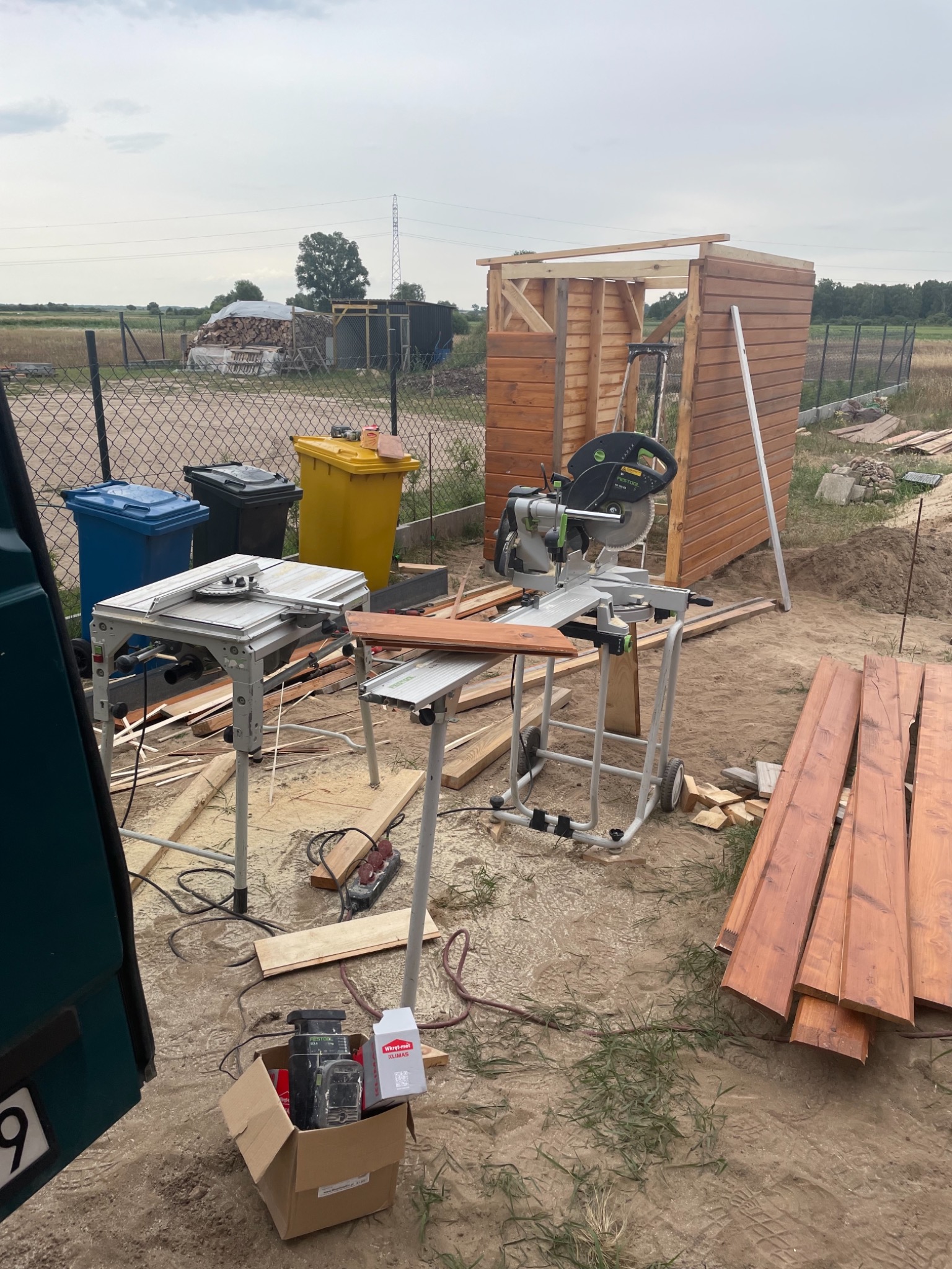 Budowa drewnianej konstrukcji szopy: szkielet z desek, narzędzia do cięcia, odpady i pojemniki na śmieci w tle.