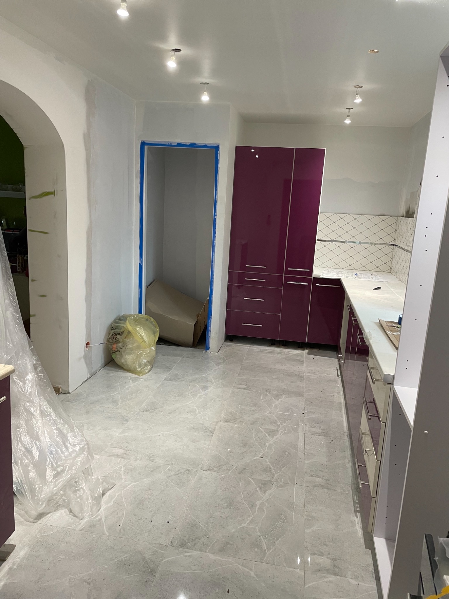Wnętrze kuchni w trakcie remontu: fioletowe szafki kuchenne, szare płytki podłogowe, ściany przygotowane do malowania, widoczny worek na śmieci i kartonowe pudło.