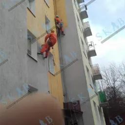 Dwóch alpinistów przemysłowych w pomarańczowych kombinezonach maluje elewację bloku mieszkalnego na żółto, zawieszeni na linach. Jeden maluje ścianę pod oknem, drugi wyżej. Na dole widoczna postać...