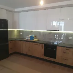 Nowoczesna kuchnia z białymi szafkami górnymi na wysoki połysk i drewnopodobnymi szafkami dolnymi, podświetlony blat z marmurowym wzorem, czarny piekarnik i lodówka, podłoga zabezpieczona kartonem.