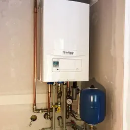 Biały piec gazowy marki Vaillant zamontowany na ścianie, z widocznymi miedzianymi rurami, manometrem i niebieskim zbiornikiem wyrównawczym, na tle surowej ściany w trakcie wykończenia.