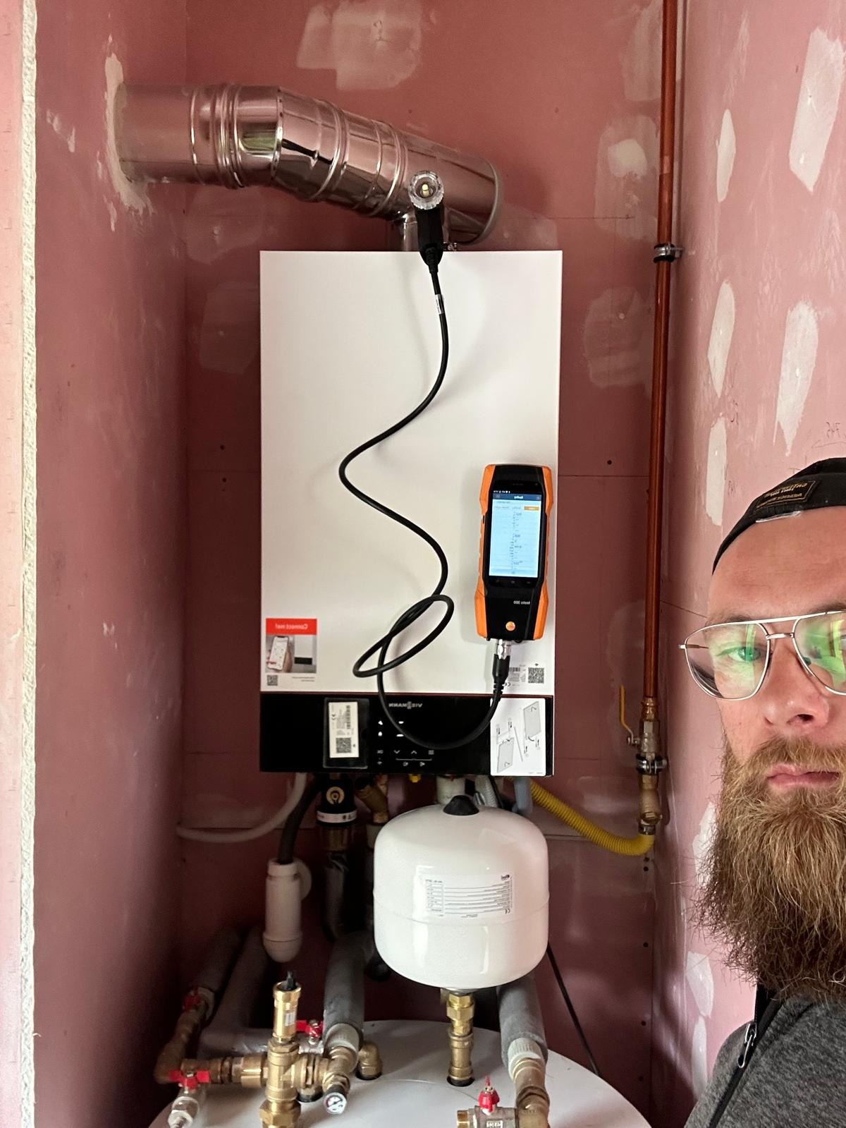 Biały piec gazowy Viessmann z podłączonym analizatorem spalin, rury miedziane i instalacja w surowym pomieszczeniu. Częściowo widoczna osoba.