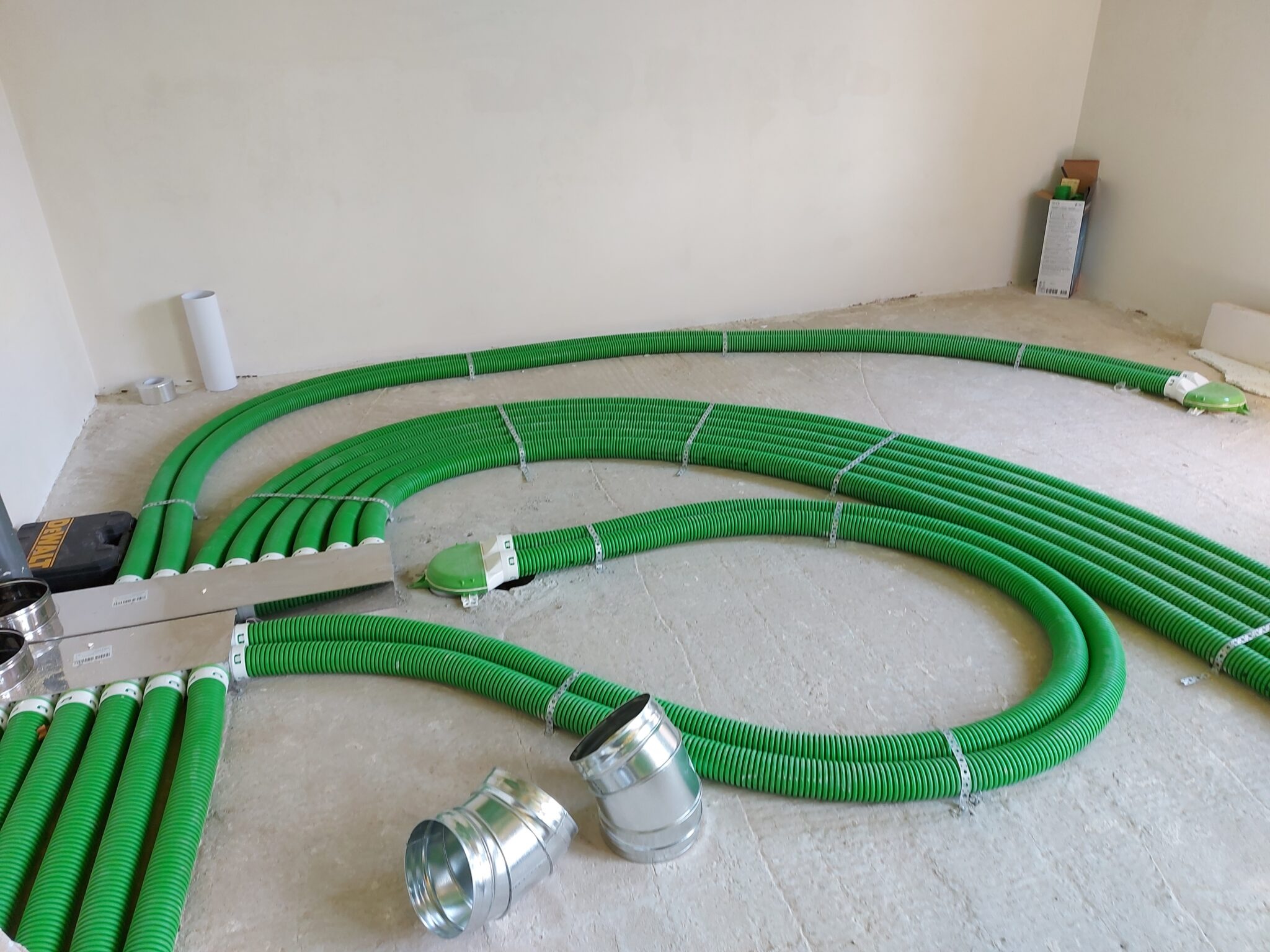 Zielone, karbowane rury wentylacyjne ułożone na podłodze w jasnym pomieszczeniu, z widocznymi elementami montażowymi i narzędziami. Instalacja w trakcie realizacji.