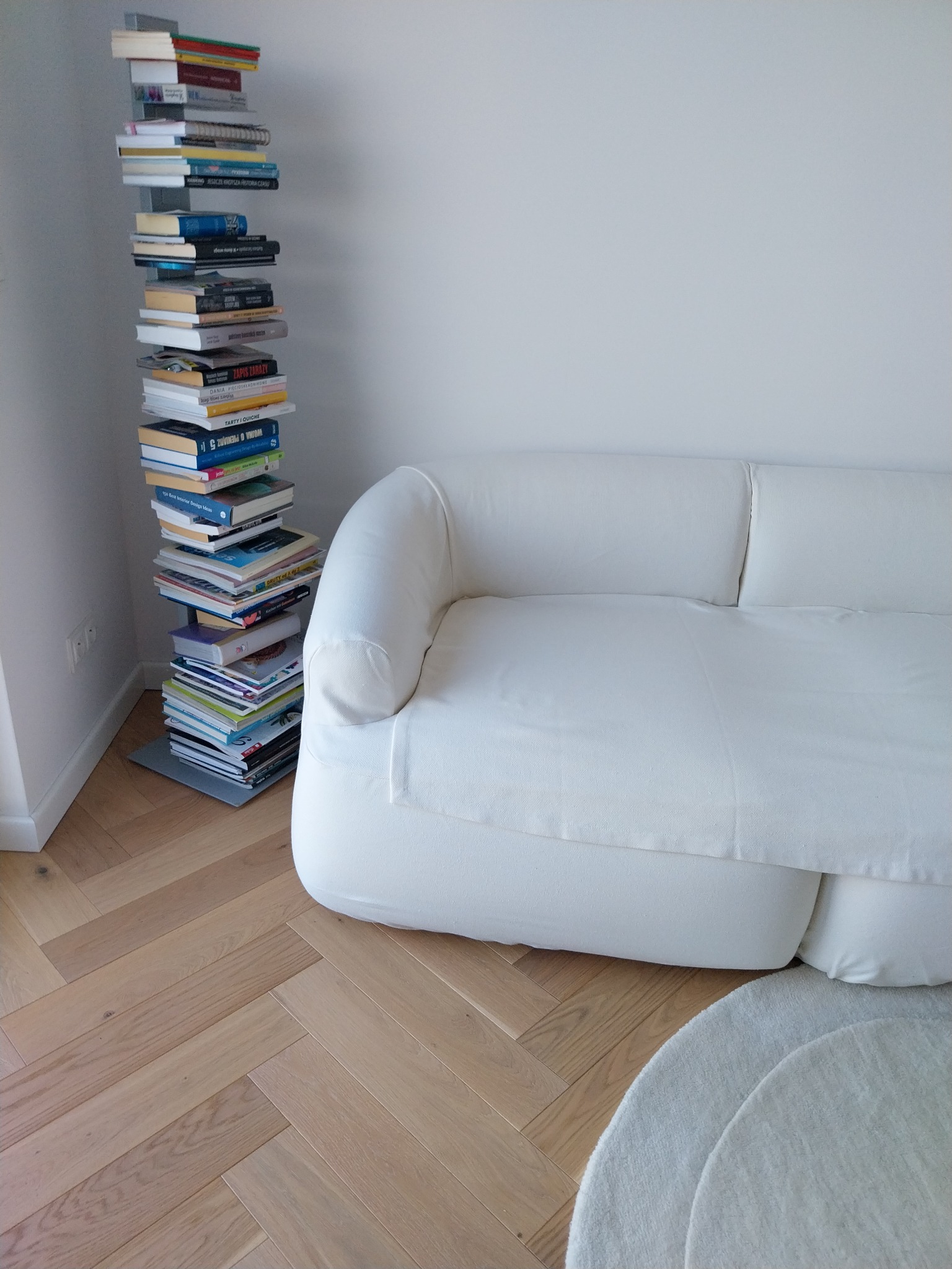 Jasny salon z białym, minimalistycznym narożnikiem i stosem książek jako nietypową kolumną, na parkiecie ułożonym w jodełkę.