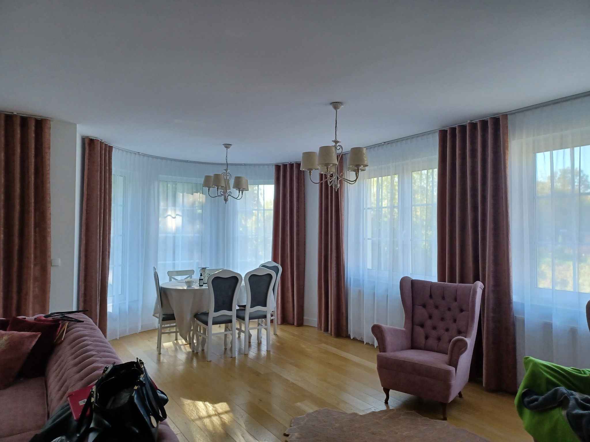 Eleganckie, brązowe zasłony na karniszu w salonie z dużymi oknami, krzesła przy stole i fotel w stylu vintage. Jasne, drewniane podłogi i subtelne oświetlenie.