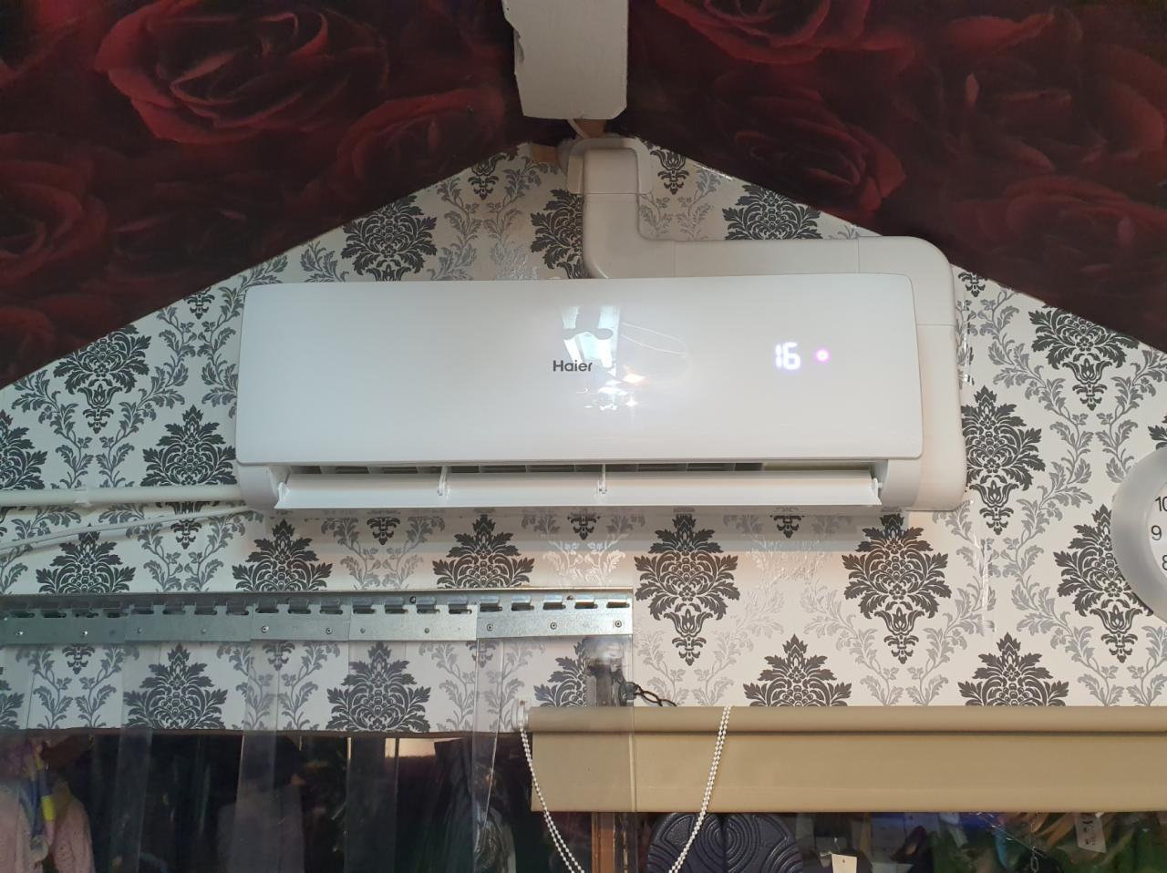Biała klimatyzacja Haier zamontowana na wzorzystej ścianie w pomieszczeniu z tapetą w róże na górze. Na wyświetlaczu klimatyzacji widoczna temperatura 16 stopni.