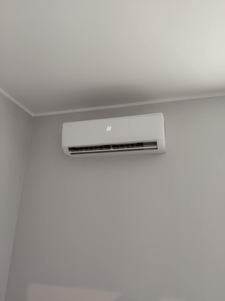 Biała klimatyzacja ścienna z wyświetlaczem temperatury (22 stopnie) zamontowana na szarej ścianie w jasnym pomieszczeniu.