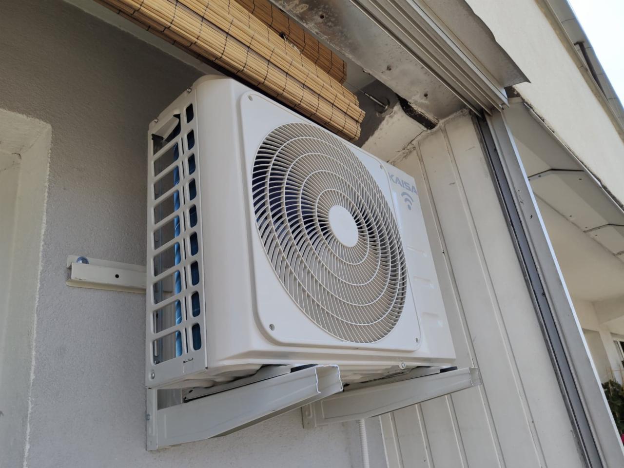 Biała jednostka zewnętrzna klimatyzatora marki KAISA zamontowana na szarej ścianie budynku pod bambusową roletą.