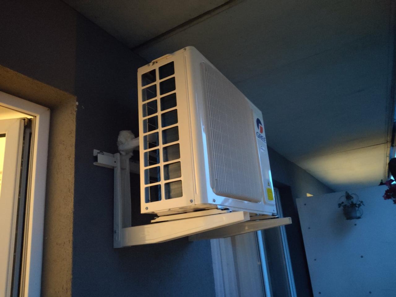 Biała jednostka zewnętrzna klimatyzatora marki Gree zamontowana na ścianie budynku z widoczną ramą okienną i fragmentem balkonu.