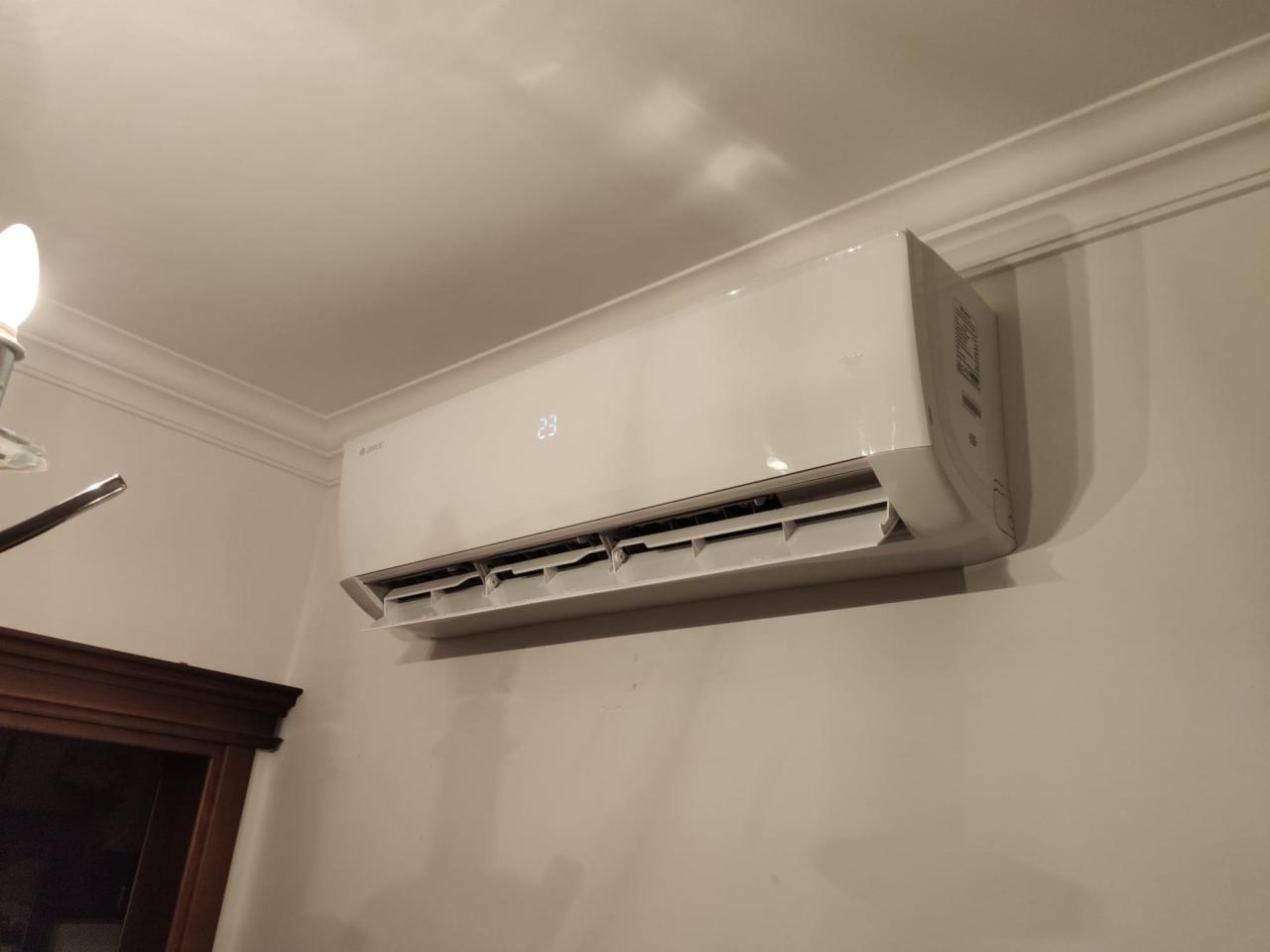 Biała klimatyzacja ścienna z wyświetlaczem temperatury 23 stopnie, zamontowana w pomieszczeniu z dekoracyjnym gzymsem sufitowym i fragmentem ciemnego obramowania drzwi.