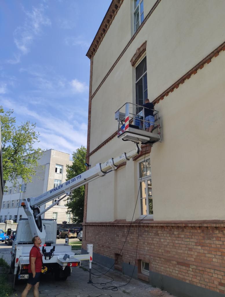 Pracownik na podnośniku koszowym przy budynku, prawdopodobnie montaż lub serwis urządzenia przy oknie na wysokości