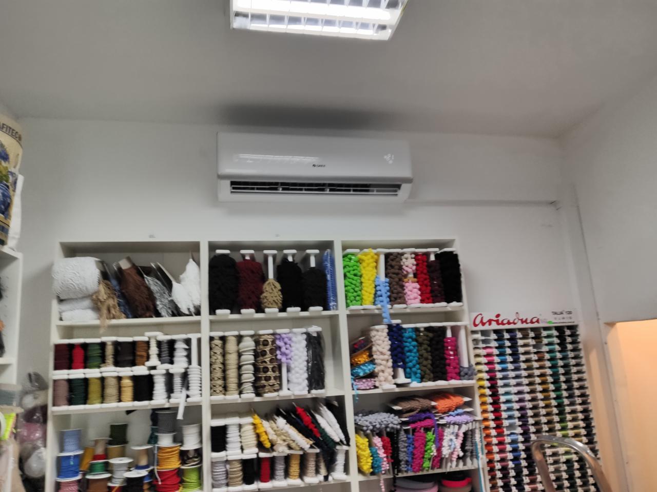 Biały klimatyzator zamontowany na ścianie nad półkami z różnorodnymi materiałami krawieckimi, takimi jak nici, koronki i wstążki w sklepie.