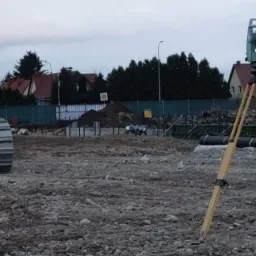 Żółta spycharka Komatsu i zielony teodolit na statywie z czerwoną skrzynką na placu budowy z fundamentami w tle, pochmurne niebo.