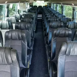 Wnętrze autobusu z rzędami czarnych skórzanych foteli, widok na przejście i okna z zielonymi drzewami w tle.
