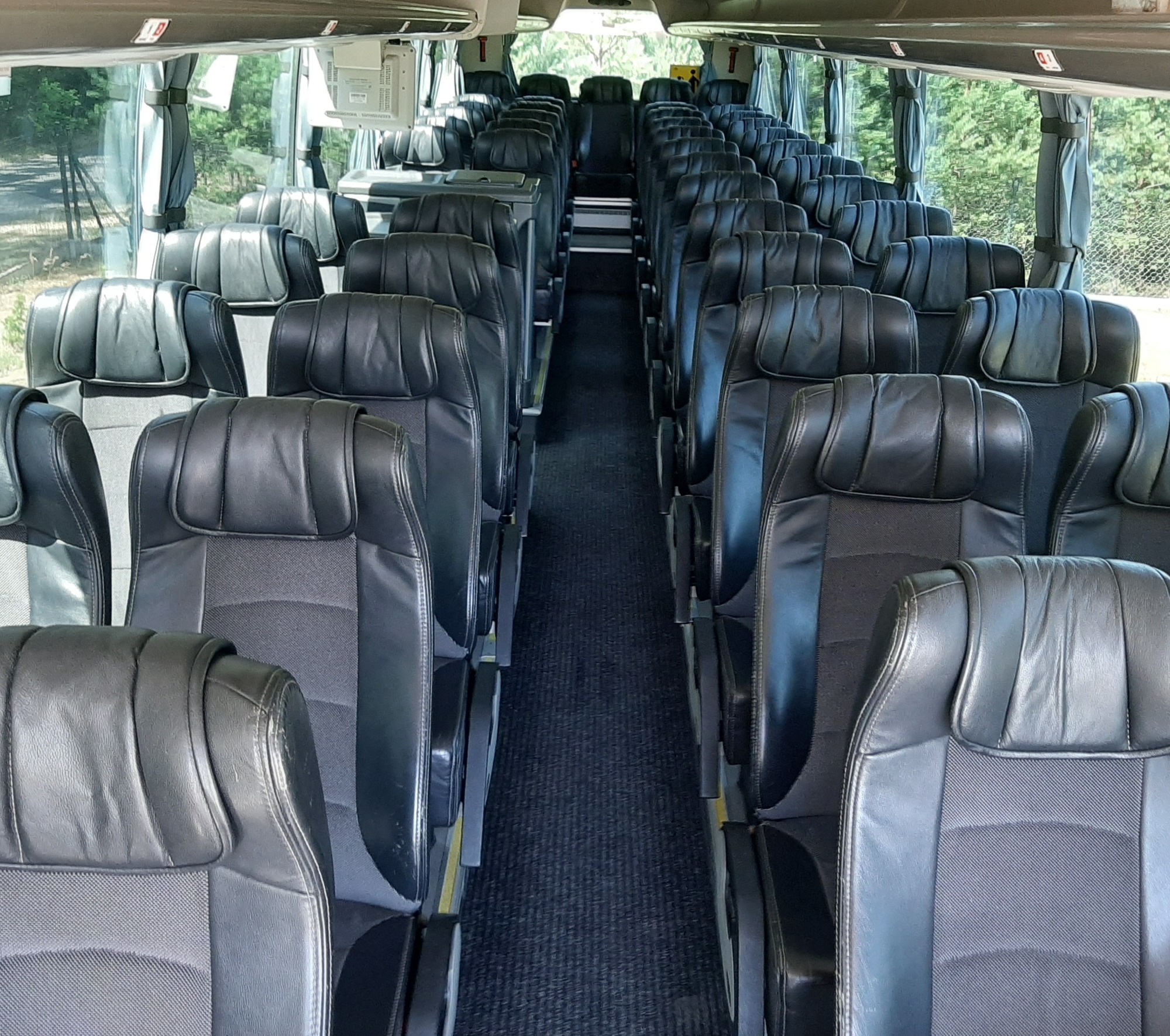 Wnętrze autobusu z rzędami czarnych skórzanych foteli, widok na przejście i okna z zielonymi drzewami w tle.