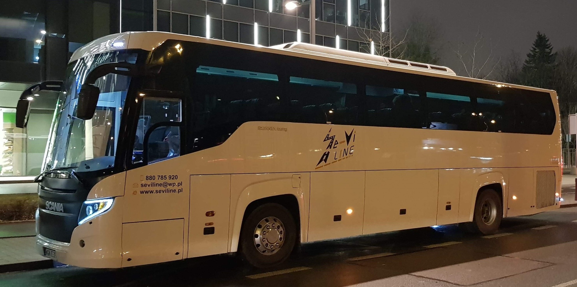Biały autokar Scania z logo Ae-Vi A Line, zaparkowany przy ulicy oświetlonej latarniami, widoczne odbicia w szybach.