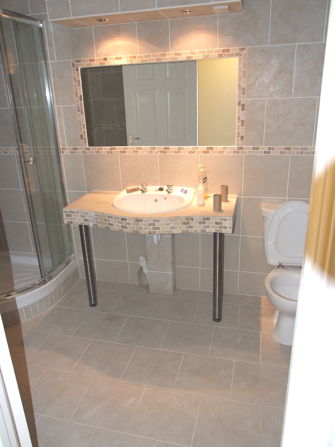 Jasna łazienka z beżowymi płytkami, kabiną prysznicową z zaokrąglonymi drzwiami, umywalką na metalowych nogach z blatem wykończonym mozaiką i toaletą z otwartą klapą.