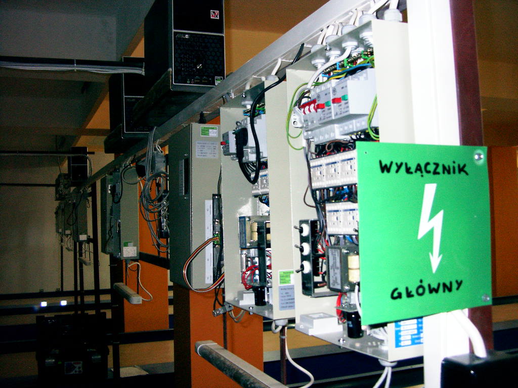 Rząd szaf sterowniczych z otwartą jedną szafą, na której widnieje zielona tabliczka z napisem 'Wyłącznik Główny' i symbolem błyskawicy, powyżej zamontowane komputery