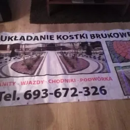 Baner reklamowy z wizualizacjami układania kostki brukowej, granitowej, wjazdów, chodników i podwórek, leżący na drewnianej podłodze, z podanym numerem telefonu kontaktowego.