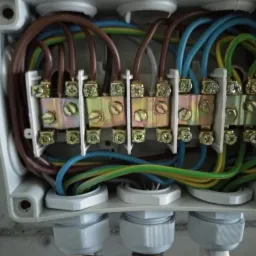 Otwarta puszka instalacyjna z widocznymi przewodami w różnych kolorach podłączonymi do listwy zaciskowej z metalowymi stykami i śrubami.