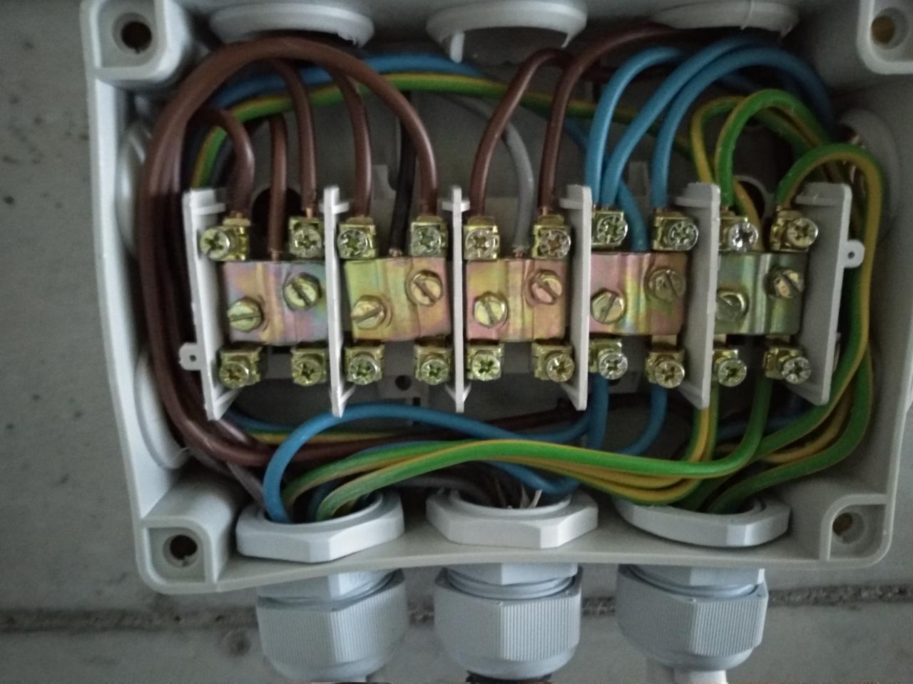 Otwarta puszka instalacyjna z widocznymi przewodami w różnych kolorach podłączonymi do listwy zaciskowej z metalowymi stykami i śrubami.