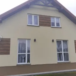 Dwukondygnacyjny dom z beżową elewacją, brązowym pasem u dołu i panelami imitującymi drewno na ścianach, widoczne białe okna z firankami i brązowy dach.