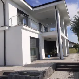 Nowoczesny, biały dom jednorodzinny z balkonem i szklanymi balustradami w Zamościu, widok z zewnątrz na świeżo pomalowaną elewację i zadbany ogr&oacute;d.