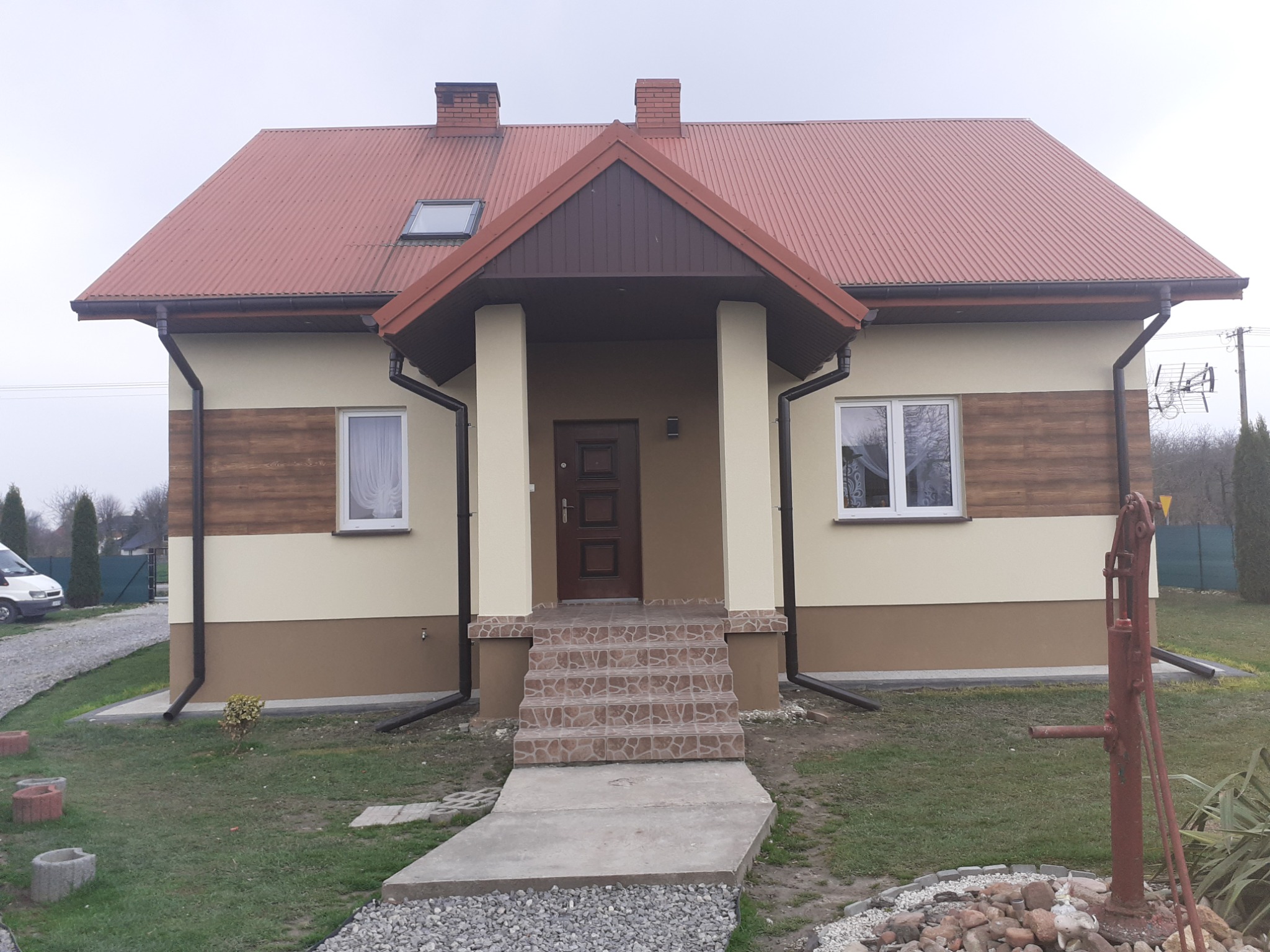 Dwukondygnacyjny dom jednorodzinny z czerwoną blachodachówką, beżowo-brązową elewacją z elementami imitacji drewna, kamiennymi schodami wejściowymi i starą, rdzewiejącą pompą wodną na pierwszym...