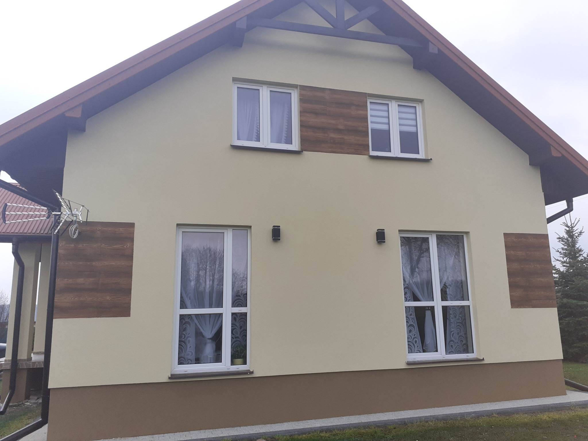 Dwukondygnacyjny dom z beżową elewacją, brązowym pasem u dołu i panelami imitującymi drewno na ścianach, widoczne białe okna z firankami i brązowy dach.
