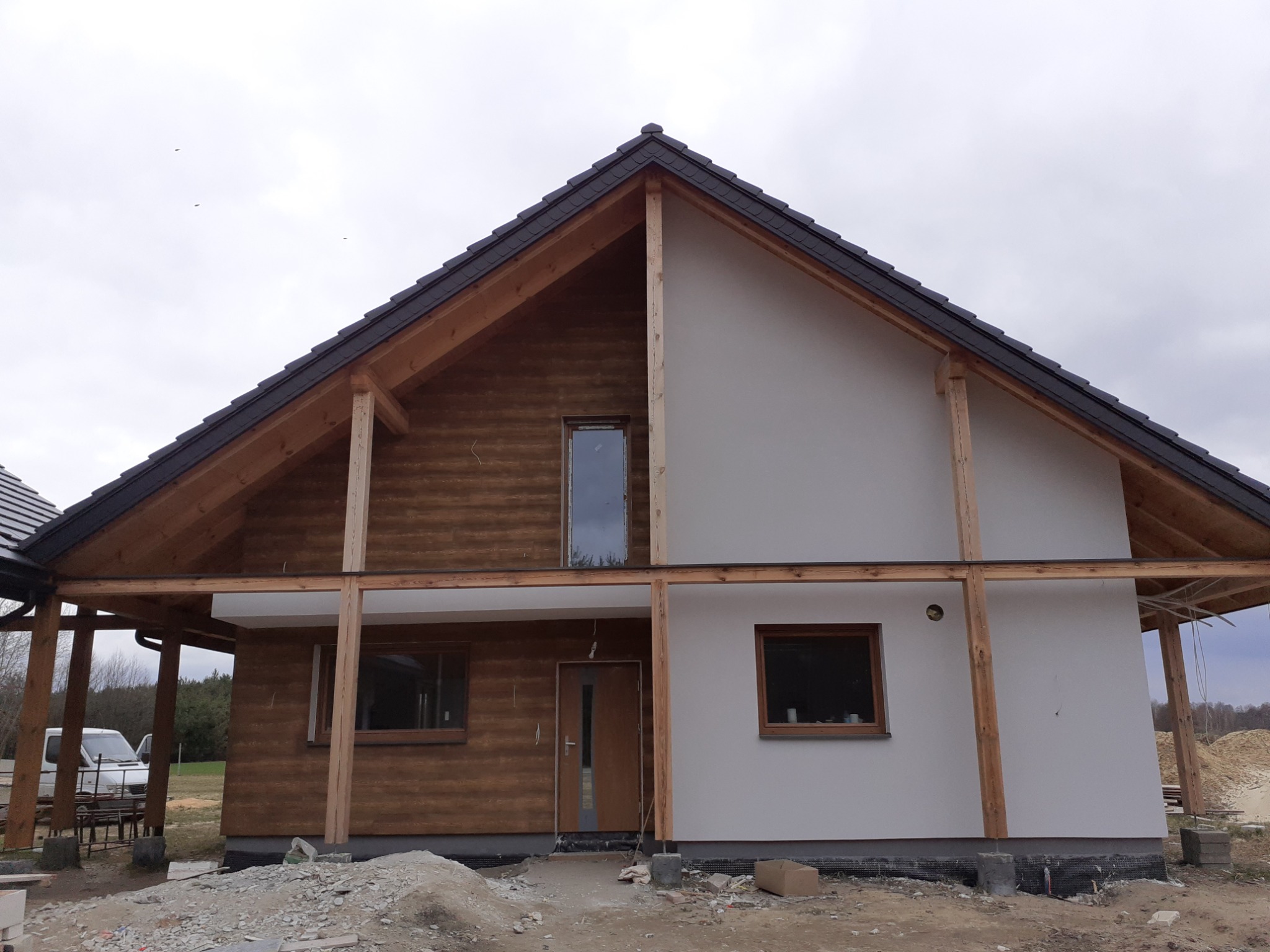 Dom jednorodzinny w trakcie prac wykończeniowych elewacji, widoczne drewniane elementy konstrukcyjne, fragment otynkowanej ściany oraz ściana wykończona drewnianą boazerią, teren wokół domu...