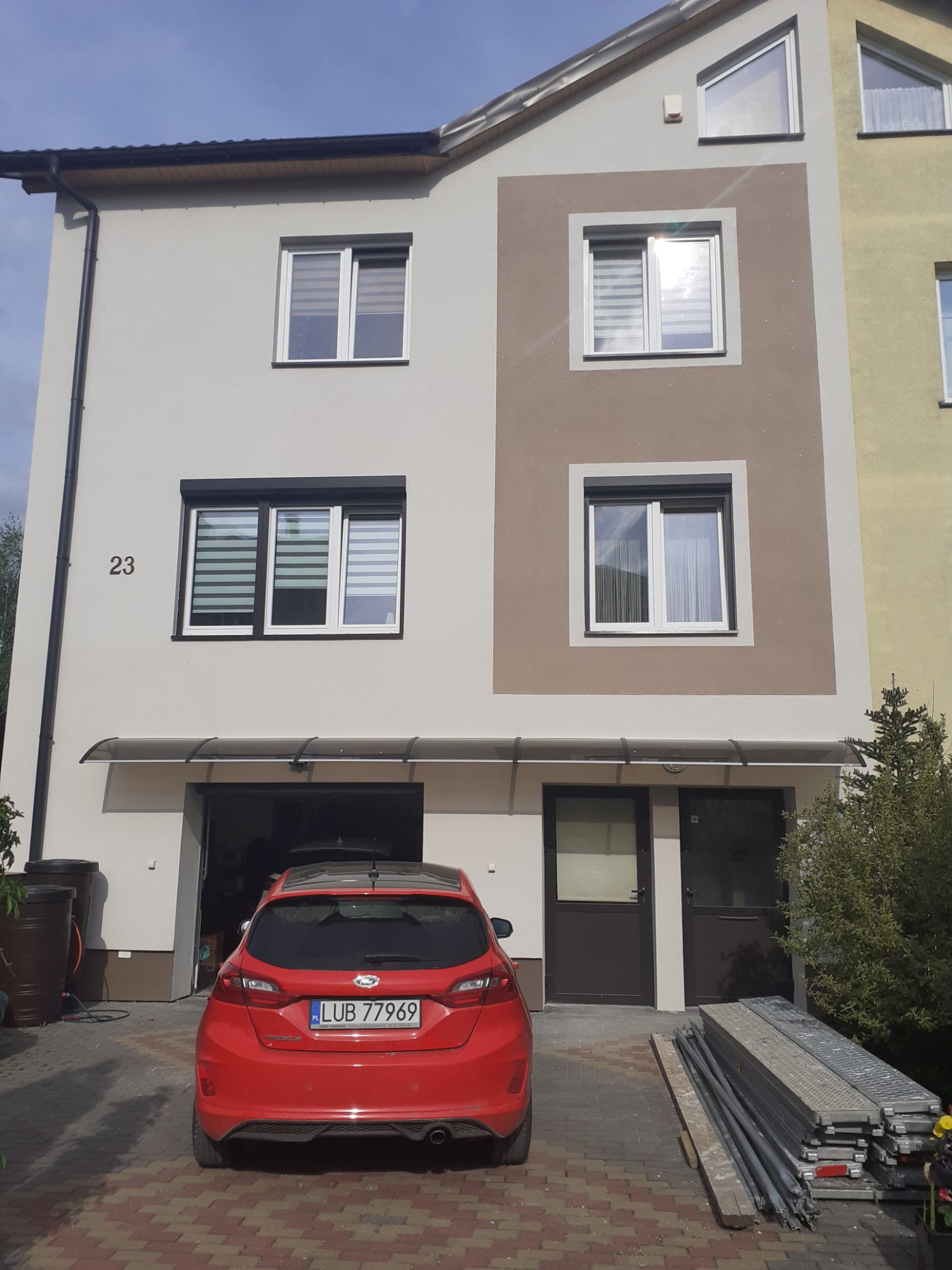 Dwukondygnacyjny dom z jasną elewacją, brązowym akcentem wokół okna na górze i numerem 23 obok okna po lewej, czerwony samochód zaparkowany pod zadaszeniem garażu, rusztowania leżące na brukowanym...