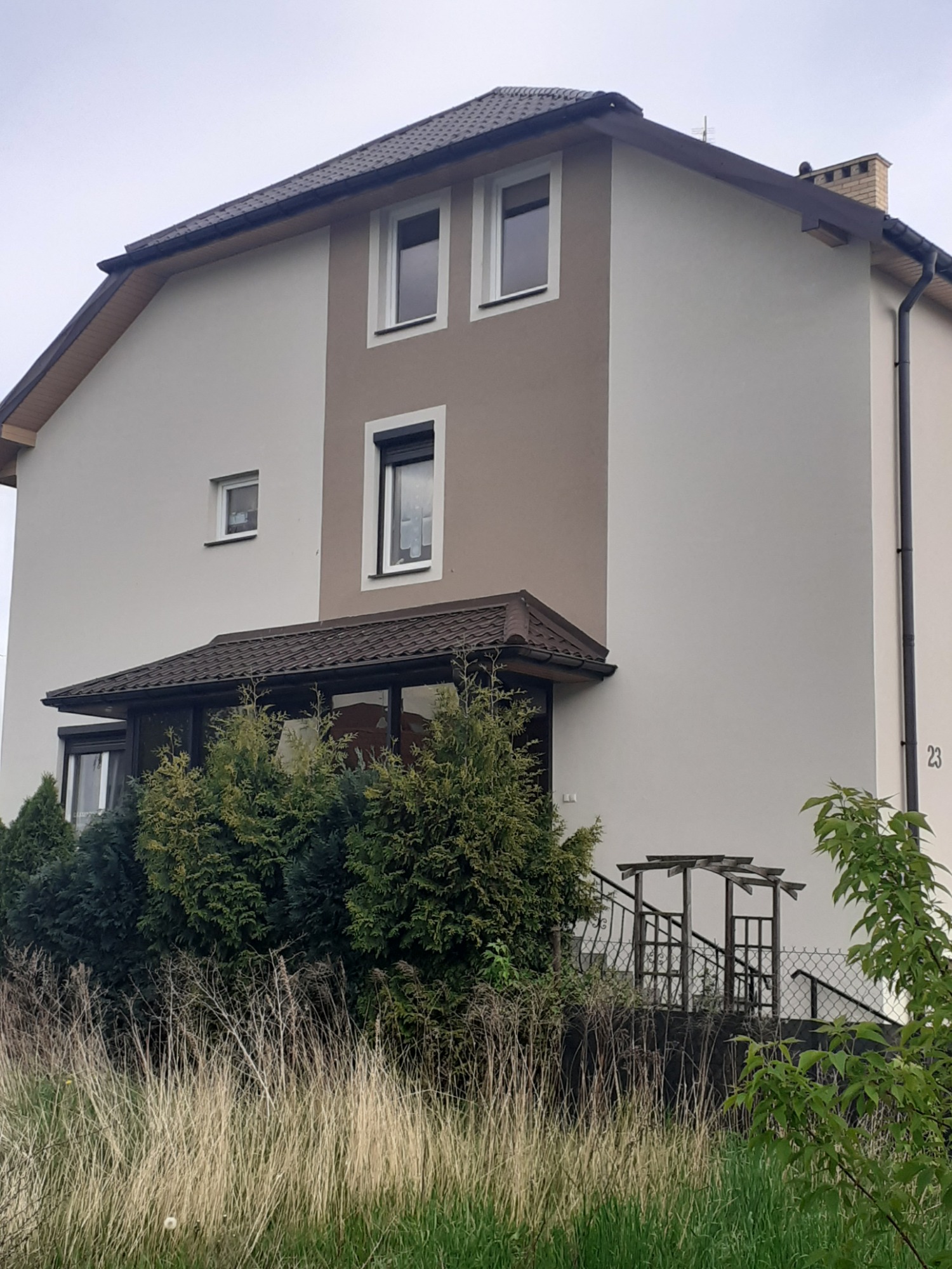 Dwukondygnacyjny dom jednorodzinny z elewacją w odcieniach beżu i szarości, z widocznym numerem domu '23' i zadaszonym wejściem porośniętym zielenią.