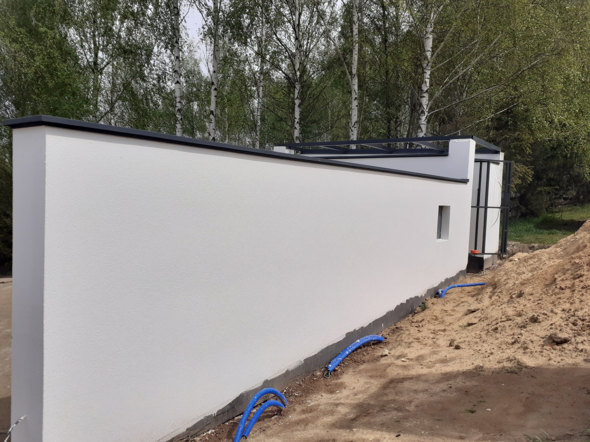 Nowoczesny, biały mur z drobną fakturą, wykończony czarnymi elementami, otoczony zielenią drzew i niebieskimi rurami na ziemi.