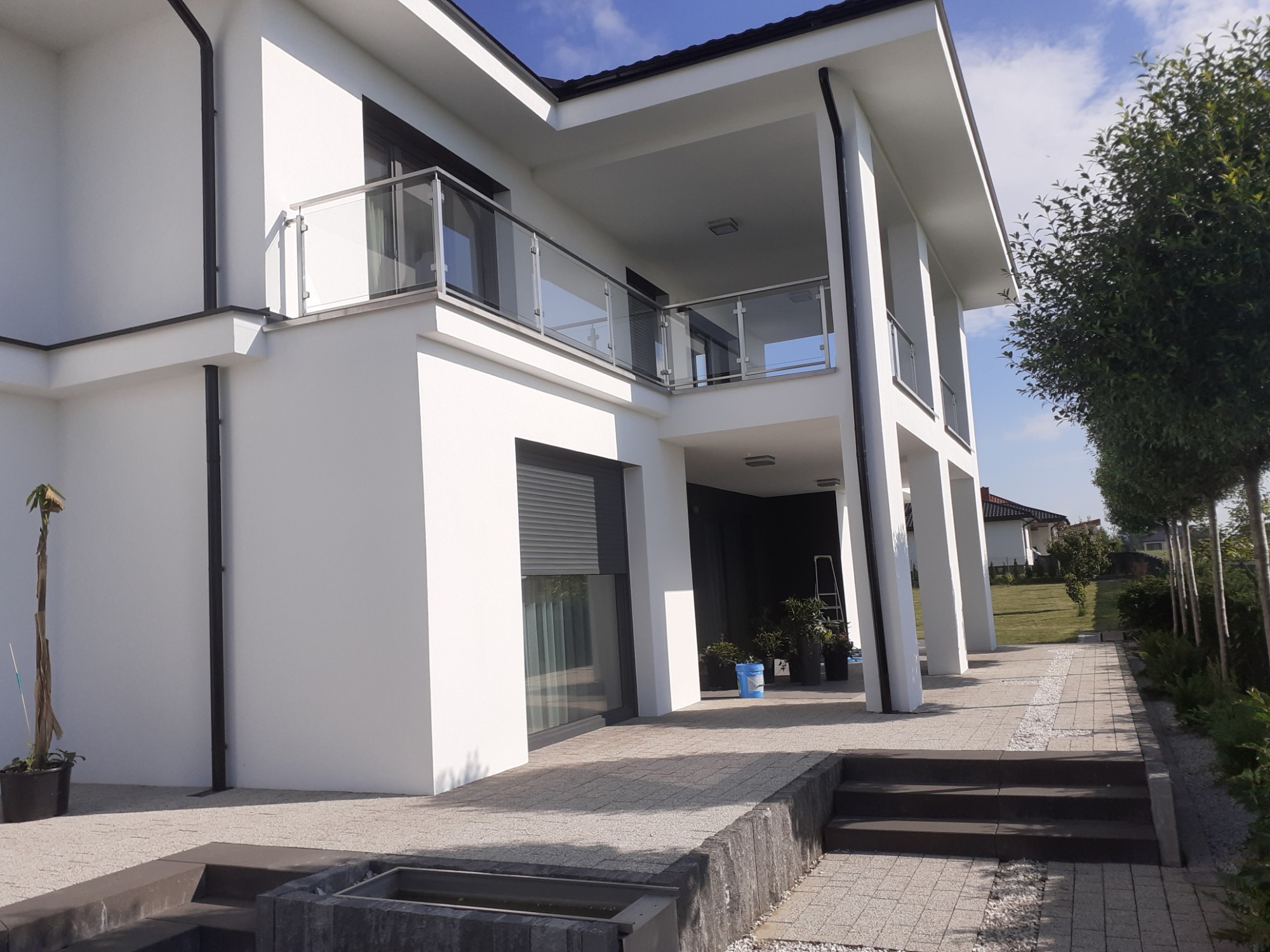 Nowoczesny, biały dom jednorodzinny z balkonem i szklanymi balustradami w Zamościu, widok z zewnątrz na świeżo pomalowaną elewację i zadbany ogród.