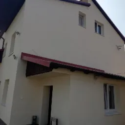 Dwupiętrowy dom jednorodzinny z jasną elewacją, czerwonymi dachówkami i szarymi schodami wejściowymi, widziany z perspektywy bocznej.