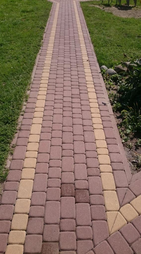 Ścieżka z kostki brukowej w odcieniach brązu i beżu, prowadząca przez trawnik w słoneczny dzień.