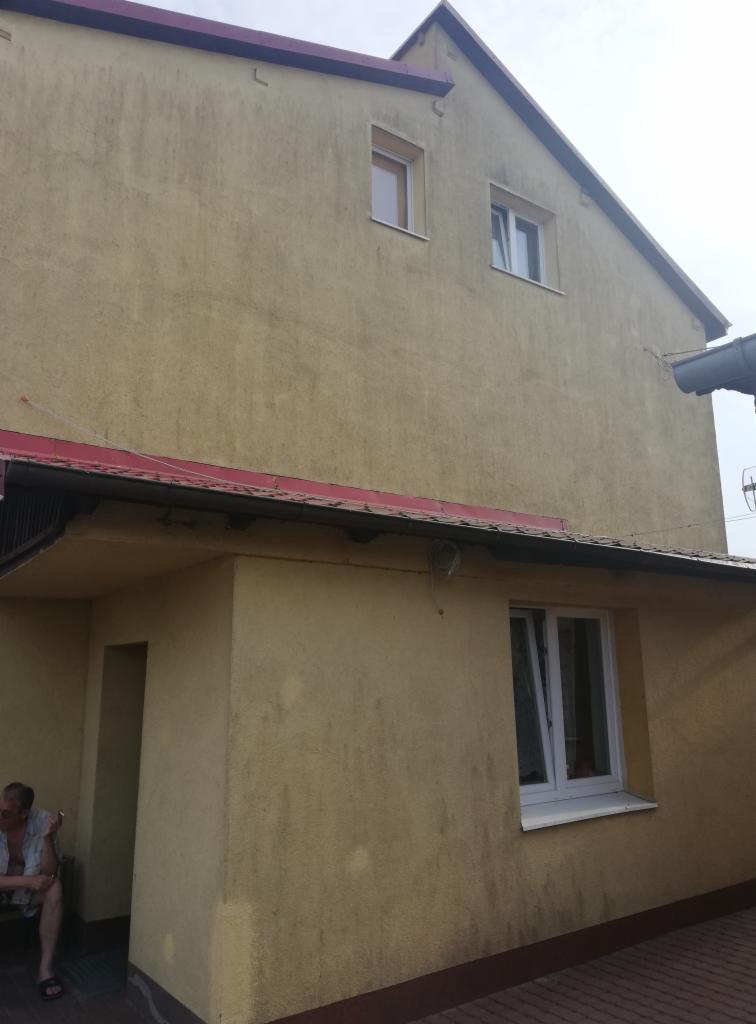 Żółta elewacja dwukondygnacyjnego budynku z czerwonym dachem, widoczne zabrudzenia i ślady po zaciekach, mężczyzna siedzący na ławce w cieniu.