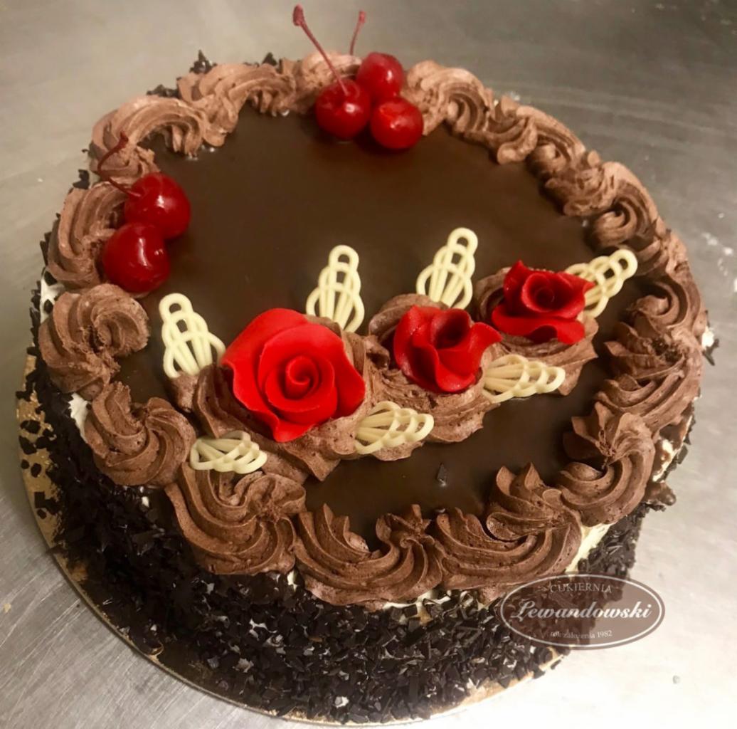Okrągły tort czekoladowy z ciemną polewą, zdobiony kremem czekoladowym, czerwonymi różami z lukru, białymi dekoracjami i wiśniami na wierzchu, na metalowym blacie.