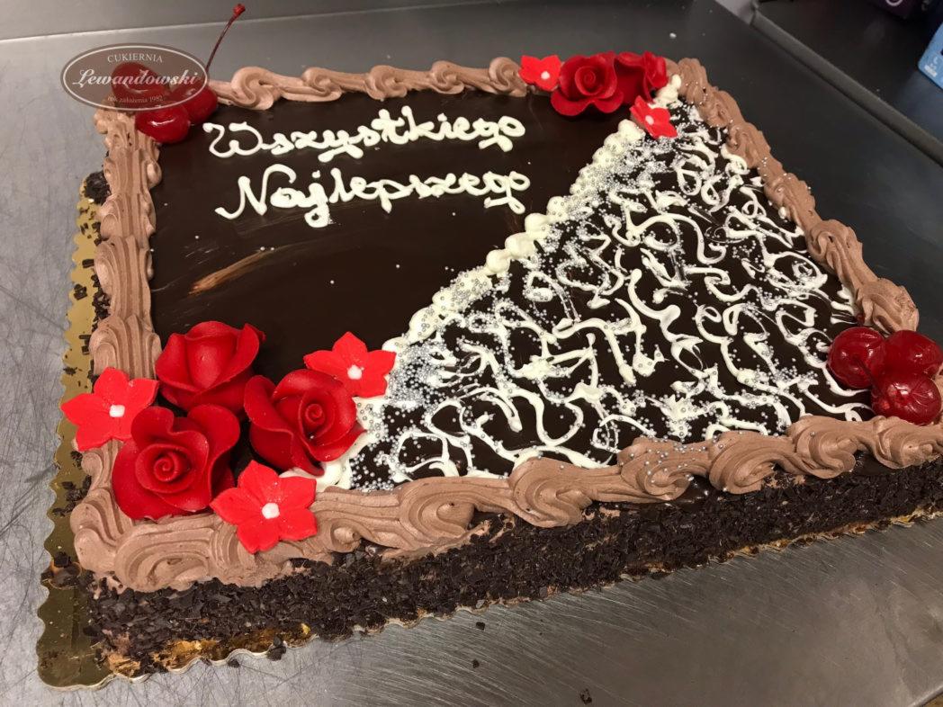 Prostokątny tort czekoladowy z napisem 'Wszystkiego Najlepszego', dekorowany czerwonymi różami, wiśniami i białymi wzorami, na metalowej powierzchni.