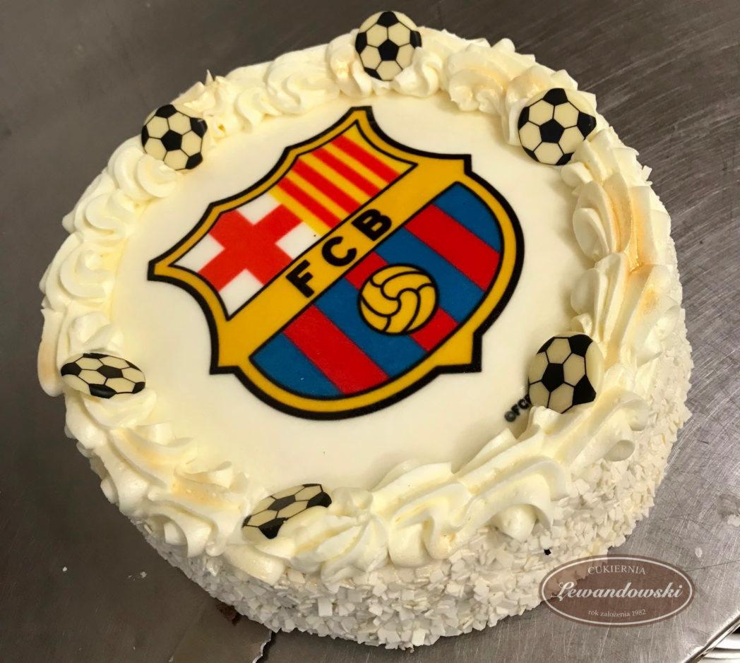 Tort z kremem, dekorowany logo FC Barcelony i czekoladowymi piłkami, na metalowym blacie.