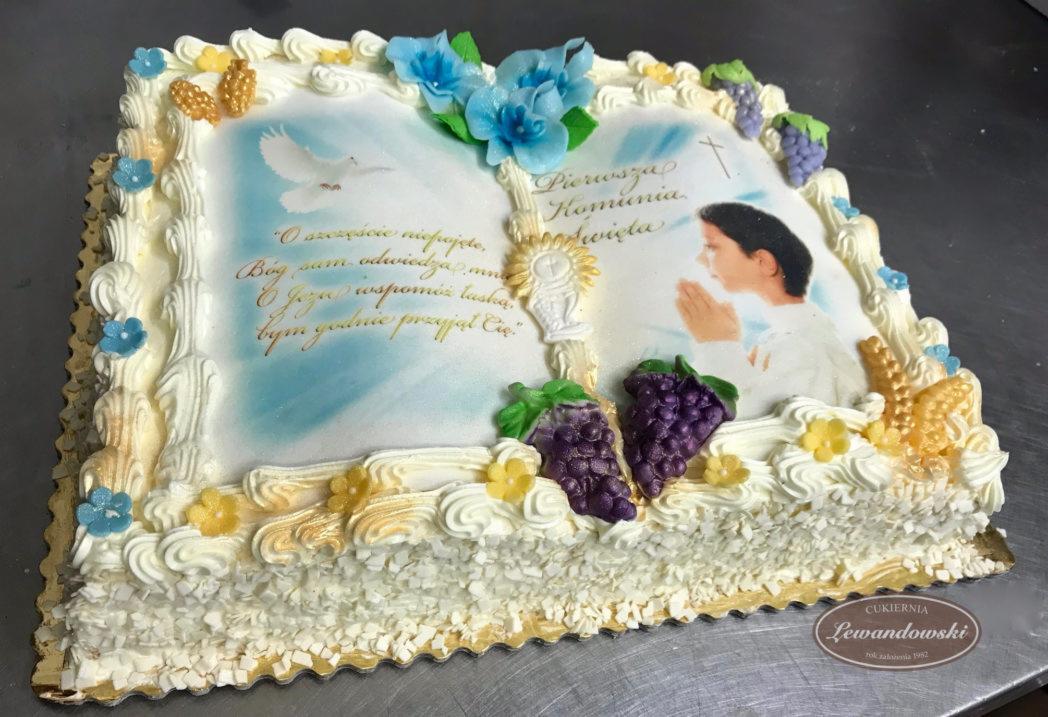 Tort prostokątny z kremem, dekorowany motywem Pierwszej Komunii Świętej, z nadrukiem wizerunku dziecka w stroju komunijnym, hostii, gołębicy i cytatem religijnym, ozdobiony kwiatami i winogronami...