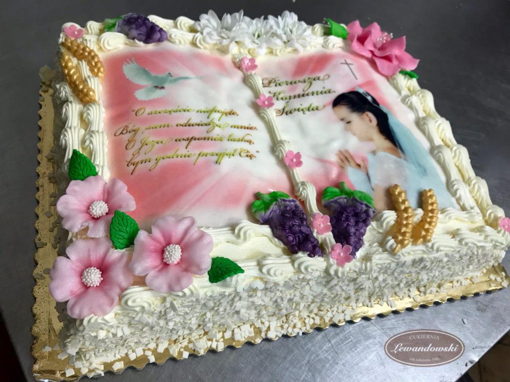 Prostokątny tort komunijny z jadalnym nadrukiem dziewczynki w welonie, gołębia i napisu religijnego, udekorowany kremem, kwiatami z lukru i imitacją winogron.