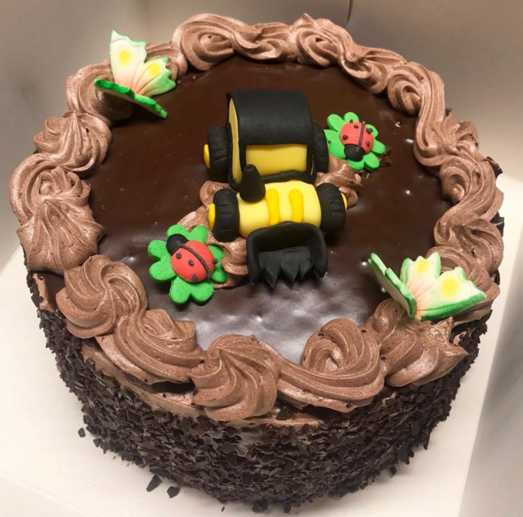Tort czekoladowy z dekoracją w postaci traktora z lukru, biedronek i motyli, otoczony kremowymi rozetkami.