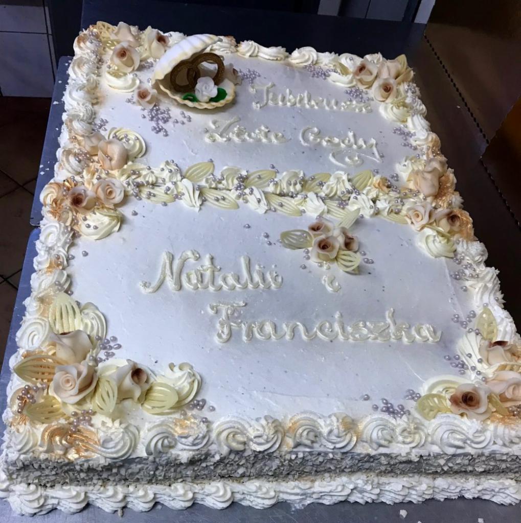 Prostokątny tort z białym kremem, dekorowany kremowymi różami, srebrnymi kuleczkami, napisem 'Jubileuszu Leśne Gody' oraz 'Natalie i Franciszka', z muszlą z pierścionkami na wierzchu.