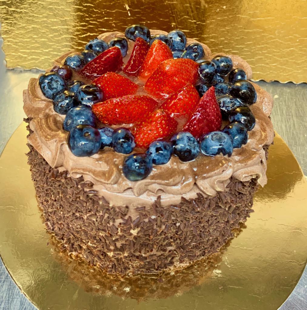 Okrągły tort czekoladowy udekorowany kremem, posypką czekoladową, świeżymi truskawkami w żelu i borówkami, umieszczony na złotej tacy.